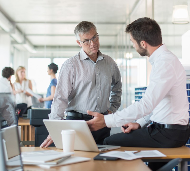 Address issues before they occur. Two small business men standing in front of laptop while team meets in background