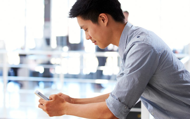 Create virtual rooms with group messaging, video calling and content and desktop sharing Businessman casually leaning over railing while using smart phone in office environment