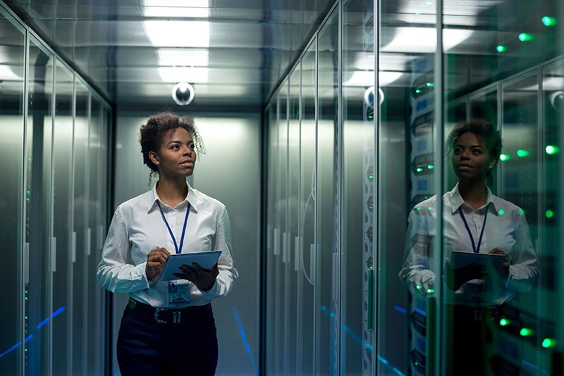 African American woman using tablet while walking in corridor of data center and checking hardware on server racks African American woman using tablet while walking in corridor of data center and checking hardware on server racks