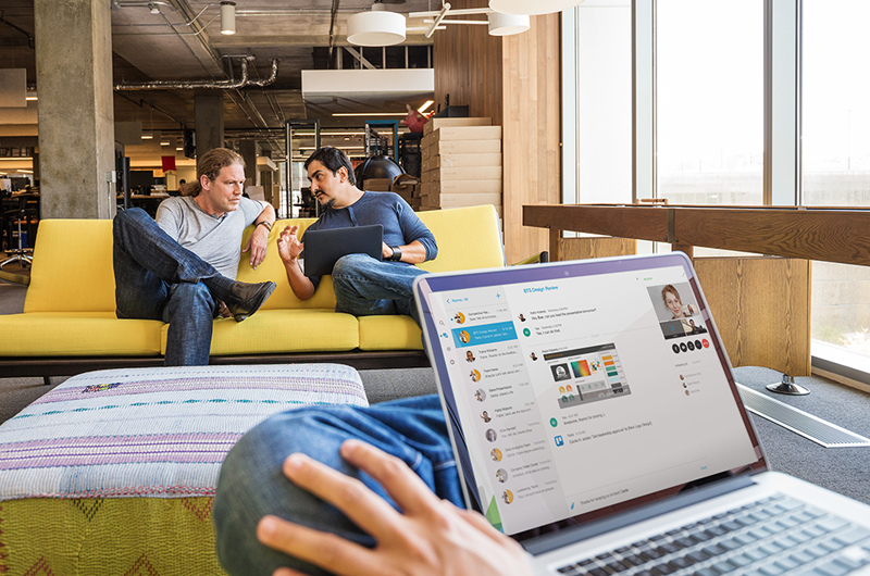 Improve sharing and collaboration Businessman videoconferences with his remote team, while two businessmen discuss plans on couch in background